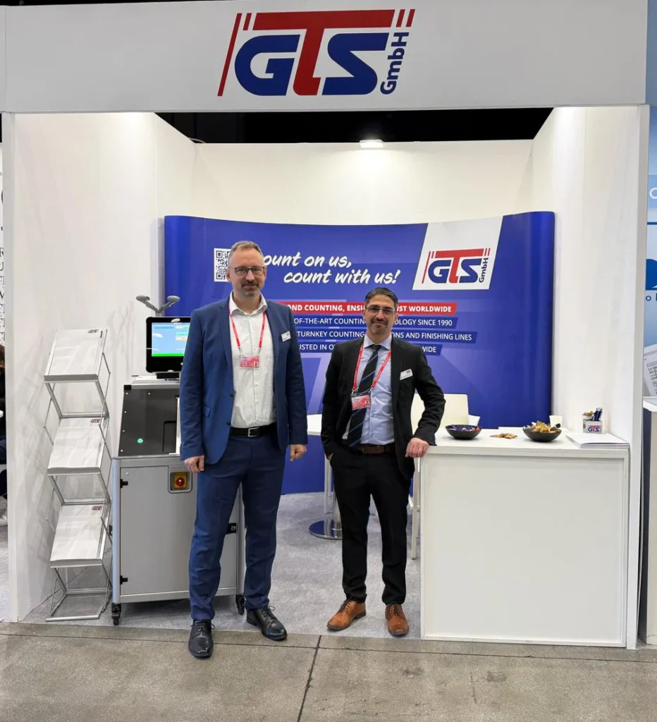 Zwei Mitarbeiter der GTS GmbH auf dem Messestand der Intergraf Messe 2025. Neben den Mitarbeitern steht eine optische Zählmaschine.