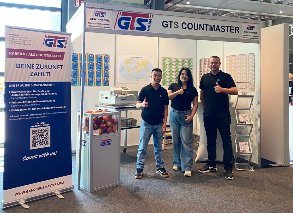 Zwei Auszubildende und ein Mitarbeiter von GTS Countmaster auf dem Messestand der Bildungsmesse Heilbronn vor einem Ausbildungsbanner mit den angebotenen Ausbildungsberufen.