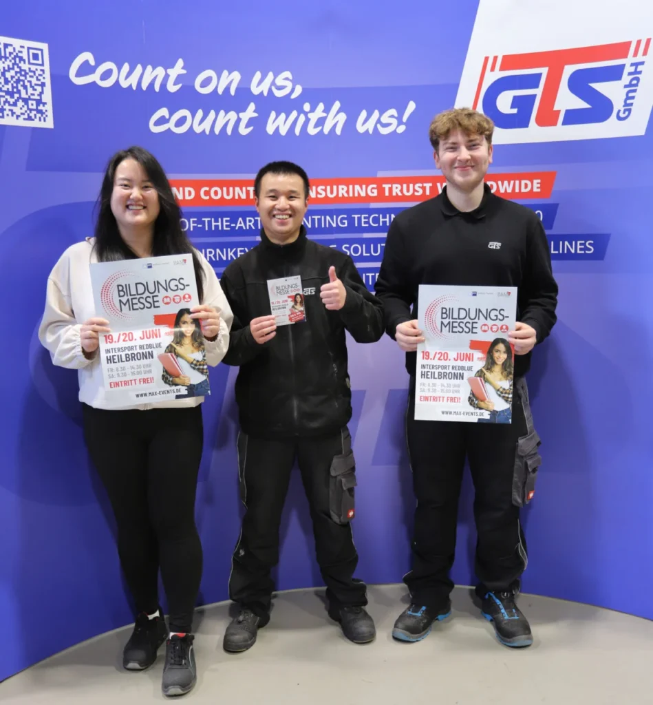 Drei Auszubildende von GTS Countmaster stehen mit Flyern der Bildungsmesse Heilbronn 2026 vor einer GTS-Werbewand