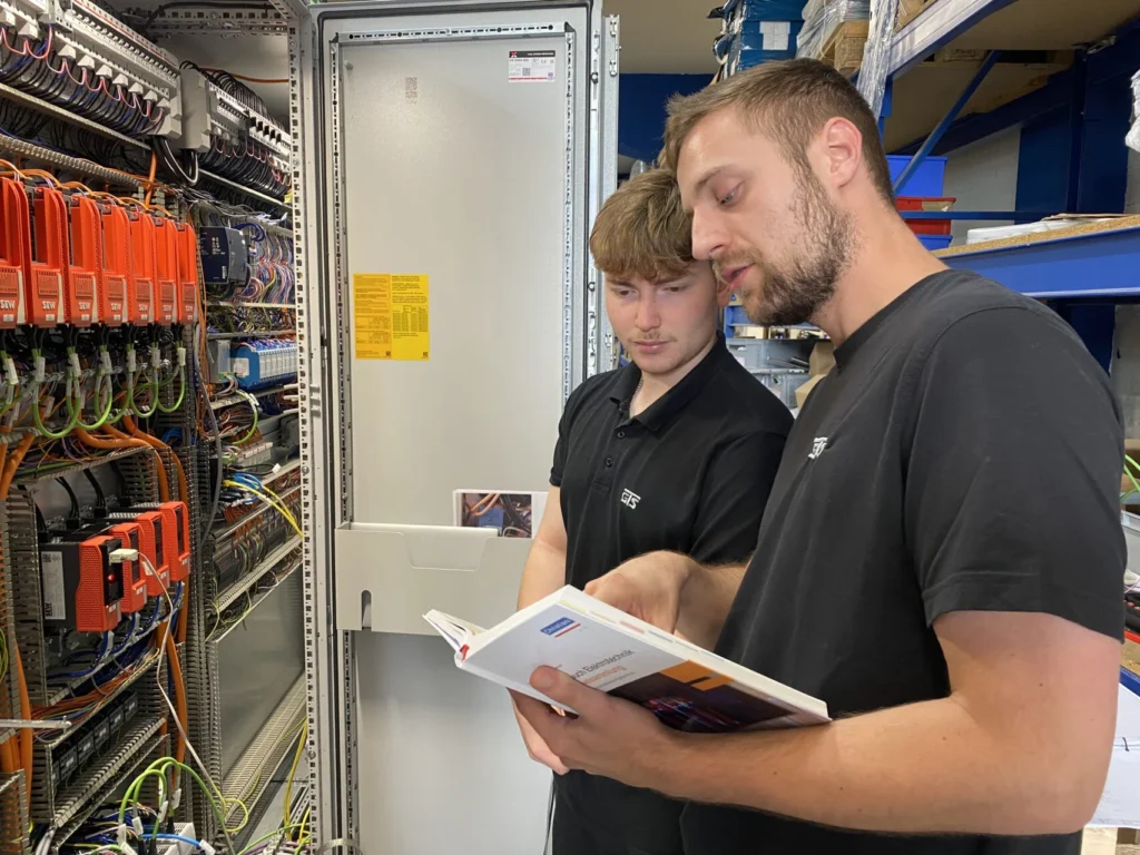 Ein Facharbeiter erklärt einem Auszubildenden zum Elektroniker für Betriebstechnik einen Schaltschrank anhand praktischer Beispiele.